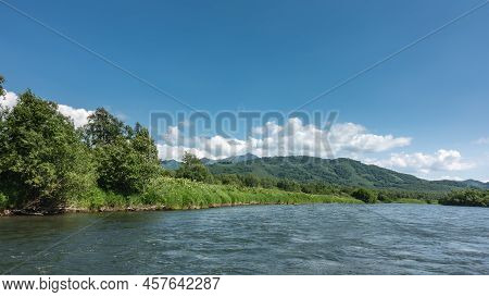 There Is Lush Vegetation On The Riverbank - Grass, Trees, Wildflowers. Mountains Against A Backgroun