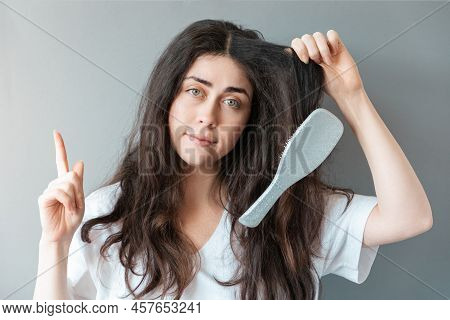 Portrait Of Young Caucasian Woman Holds A Strand Of Her Hair With A Comb Stuck In It. Gray Backgroun