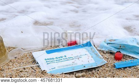 Close-up Of Used Reusable Face Mask With Plastic And Other Trash On The Beach. Sea Waves Washing Up 