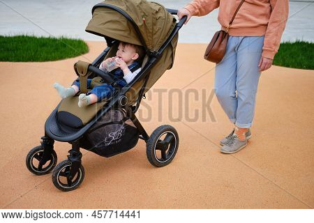 Woman Mother Walks With Toddler Baby Boy Sitting In A Stroller On The Playground Of The Park. Child 
