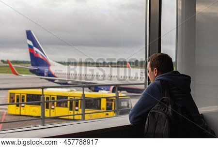 Guy Stands Near Big Window At Airport In Cloudy Weather. He Looks Outside And Watches The Traffic. J