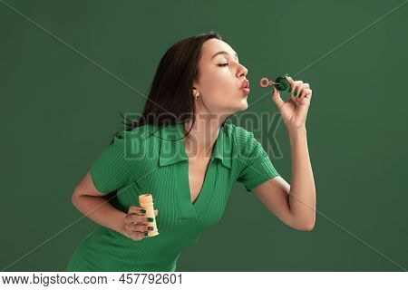 Portrait Of Beautiful Young Girl Posing, Blowing Soap Bubbles Isolated Over Green Studio Background