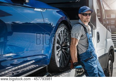 Caucasian Towing Truck Driver In His 40s Staying Next To His Vehicle. Repossessed Car On The Truck.