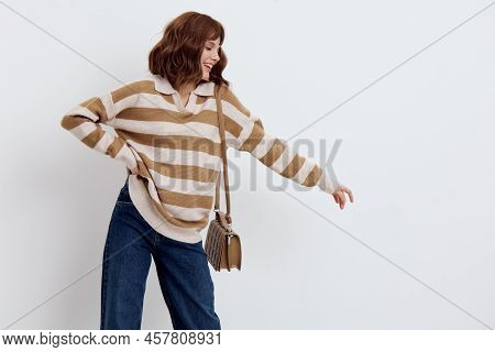 A Joyful Woman Stands On A White Background In A Striped Sweater And With A Bag On Her Shoulder Put 