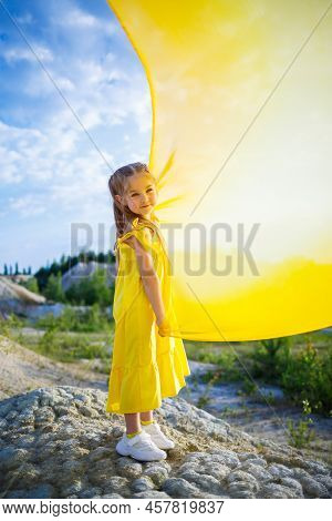 A Small 6-7 Years Old Ukrainian Girl In A Yellow Dress With Wings In Yellow Fabric By The Lake. Blue
