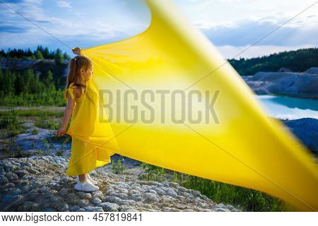 A Small 6-7 Years Old Ukrainian Girl In A Yellow Dress With Wings In Yellow Fabric By The Lake. Blue