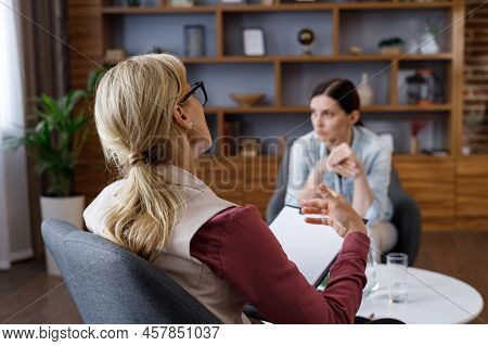 Over Shoulder View Of Female Psychologist Sitting In Armchair, Talking With Upset Woman Patient. Psy