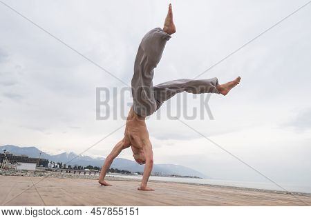 Shirtless Caucasian Man Doing Backflip On Pebble Beach.