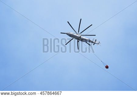 Helicopter Going To Fire Area To Extinguish Forest Fire. Helicopter With A Water Bucket On A Blue Sk