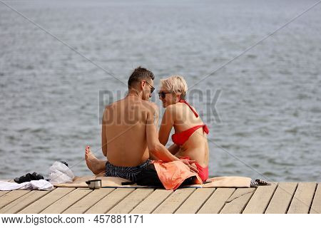 Moscow, Russia - July 2022: Happy Couple Sunbathing On A River Beach Sitting On Wooden Pier, Summer 