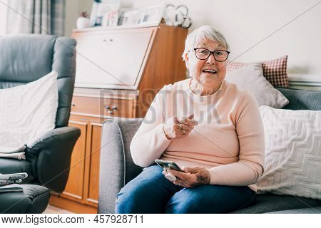 Portrait Of Happy Laughing Senior Woman Using Phone While Sitting Onon The Sofa At Home. Mature Olde
