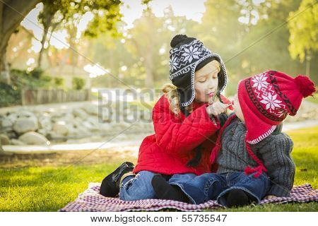 Little Girl with Her Baby Brother Wearing Winter Coats and Hats Sharing a Lollipop Outdoors at the Park. 