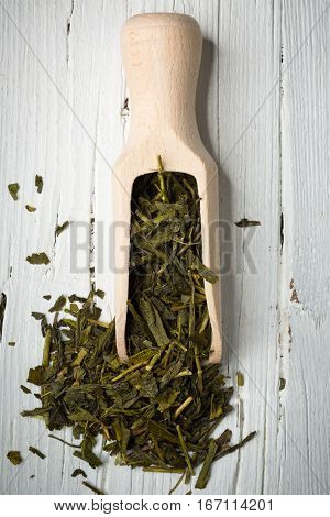 Dried green tea leaves with scoop on white wooden background view from above flat lay.