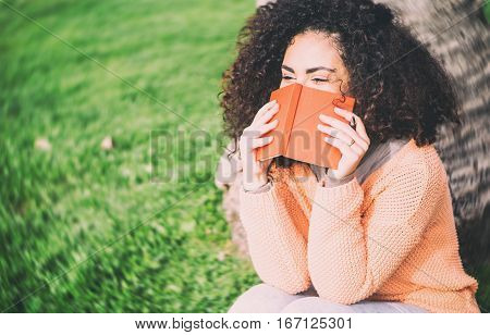 Brunette student girl with curly hair covering her face with her diary looking embarrassed sitting against a tree on a grass of park - Young woman writing on her copybook laughing outdoor