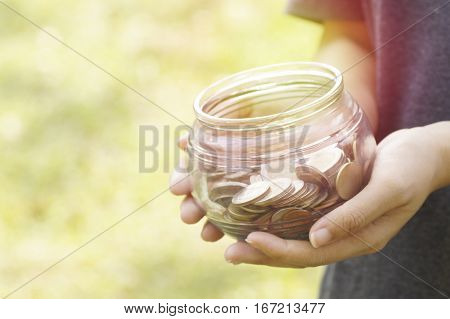 young girl hold glass bottle of coins to save money and donation concept in blur green gardent
