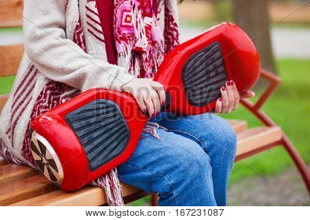 Girl in boho style clothes sitting on a bench holding modern electric mini segway or hover board scooter in hands. Trending new transportation technology that produces no air pollution to the atmosphere. Unrecognizable model and close up on the device