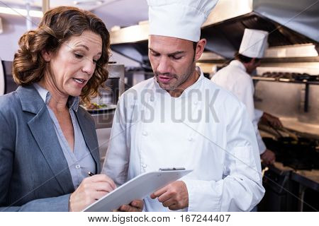 Female restaurant manager writing on clipboard while interacting to head chef in commercial kitchen