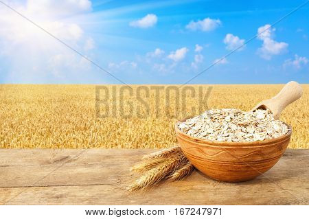 Oat flakes in bowl. Ears of oats and oatmeal in bowl on table with field on the background. Ripe wheat field, blue sky, sun. Uncooked porridge