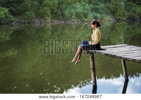 Dreaming girl sitting on bridge with forest and river on the background. Lonely sad woman by the lake. Beautiful woman in elegant vintage blouse, skirt, floppy hat. Natural sensual woman outdoors