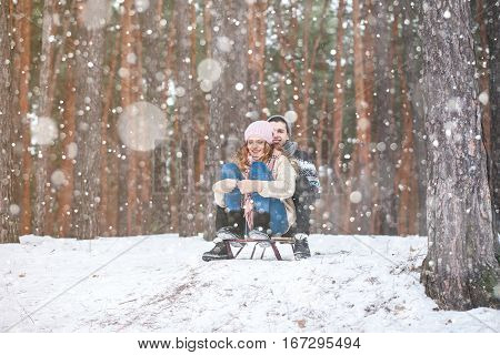 Young couple in winter forest sledding, laughing during snowfall