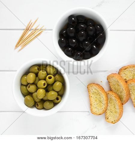 Pitted black and green olives in bowls with crostini on the side photographed overhead on white wood with natural light