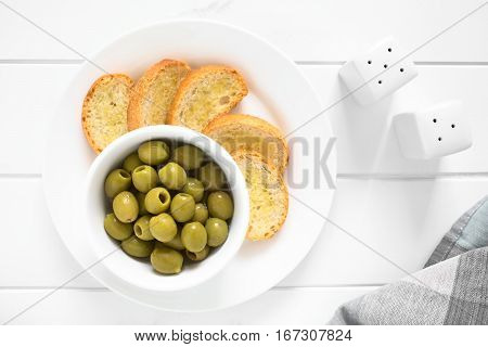 Pitted green olives in bowl with crostini on the side photographed overhead on white wood with natural light