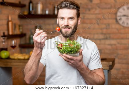 Handsome bearded man in white t-shirt eating salad with tomatoes in the kitchen. Healthy and vegan food concept