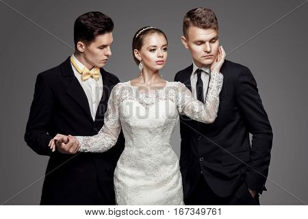 Two handsome men in suits and beautiful woman in white bridal dress. Studio, trendy, strict look.