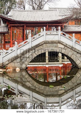 A traditional Chinese bridge reflects in a calm pond in a memorial park in the old city of Dali China.