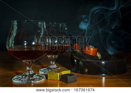 Scotch drink and tobacco pipe with smoke on black background. Two old fashioned whiskey glass and chocolate piece. Unhealthy still life or bad habits concept. Clear brandy or bourbon snifter.