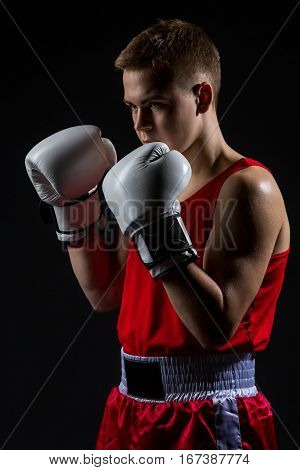 Young handsome boxer sportsman in red boxer suit and white gloves standing on black backgound. Copy space.