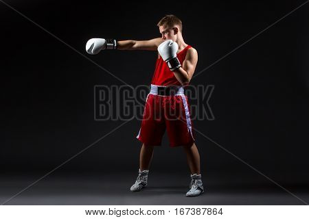 Young handsome boxer sportsman in red boxer suit and white gloves standing on black backgound. Copy space.