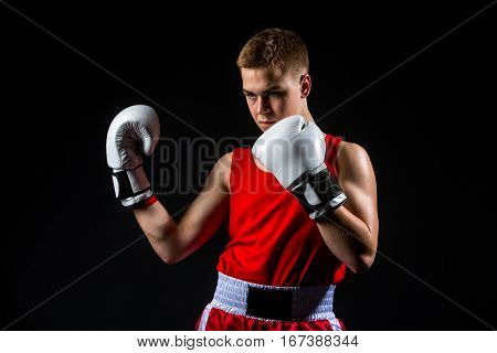 Young handsome boxer sportsman in red boxer suit and white gloves standing on black backgound. Copy space.