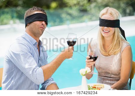 Blindfolded couple holding red wine while sitting at poolside