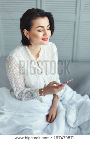 Pretty woman smiling in the morning reading the messages on the phone she sits on the bed.