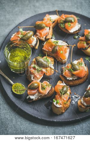 Homemade salmon crostini with cream-cheese, watercress, capers and pesto suace in round black slate stone plate over grey background, selective focus