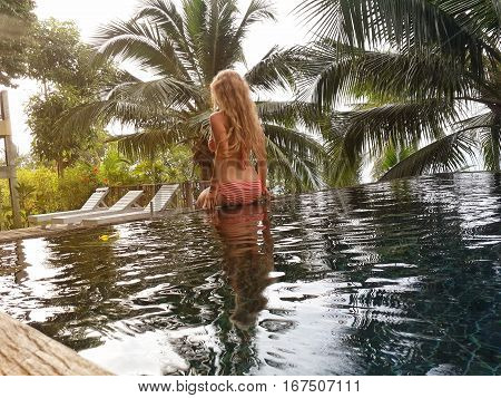 Blonde girl sits on the edge of the pool in the jungle with palm trees. Lifestyle concept