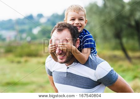Boy closes hands eyes father. Happy little boy enjoying with riding on father's back. Happy family portrait. Laughing dad with little boy enjoying nature together.