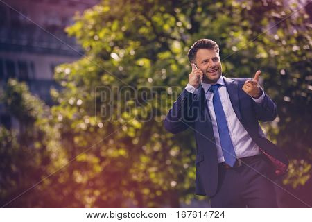 Cheerful businessman gesturing while talking on mobile phone against tree