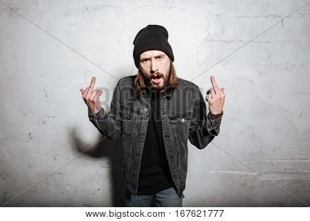 Portrait of a hipster man in hat showing fuck you sign and looking at camera isolated over wall background