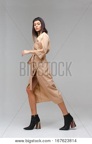 Young beautiful woman in studio shows that she is making steps. She has long light brown cloak and black boots. She is posing and playing role go away