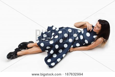 Beautiful model laying on the white floor in studio with wwhite isolated background. She is looking aside and wearing long blue dress with polka dots and big black boots