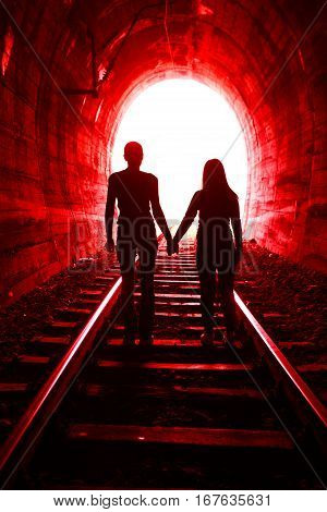 Couple walking hand in hand along the rails through a railway tunnel towards the bright at the end they appear as silhouettes against the light