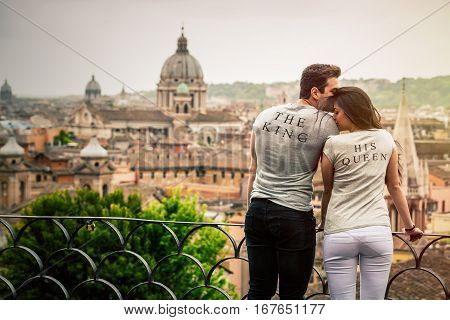 Romantic couple in Rome, Italy. Two young people, a men and a women are kissing and loving. On a terrace with railing overlooking Rome. Wearing a gray T-shirt with the words The King and His Queen.