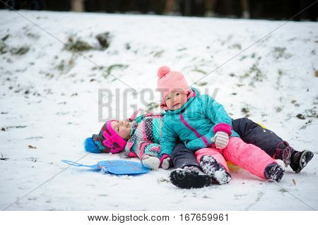 Two little girls in bright ski suits moved out with a snow slide. Children lie on snow and smile. They have rosy cheeks. Girls are glad to winter and snow.