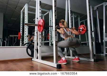 Young sexy blonde european girl in the gym doing squat with barbell in front of the mirror. She is dressed in sports clothes white top, gray leggings, orange sneakers