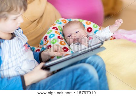 Preschool kid boy playing games tablet computer during babysitting. Cute newborn baby looking on brother. Adorable kids and children at home.