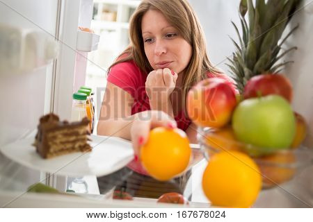 Attractive  female on diet in dilemma whether to eat piece of chocolate cake or orange from fridge