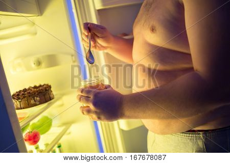Close up of fat male eat at night jam from jar with big spoon from fridge