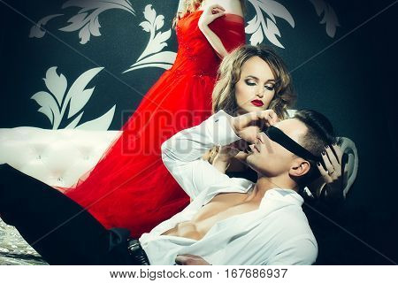 Sexy man in unbottoned shirt with tied eyes. Pretty girl in gorgeous dress tied the man's eyes with black stripe